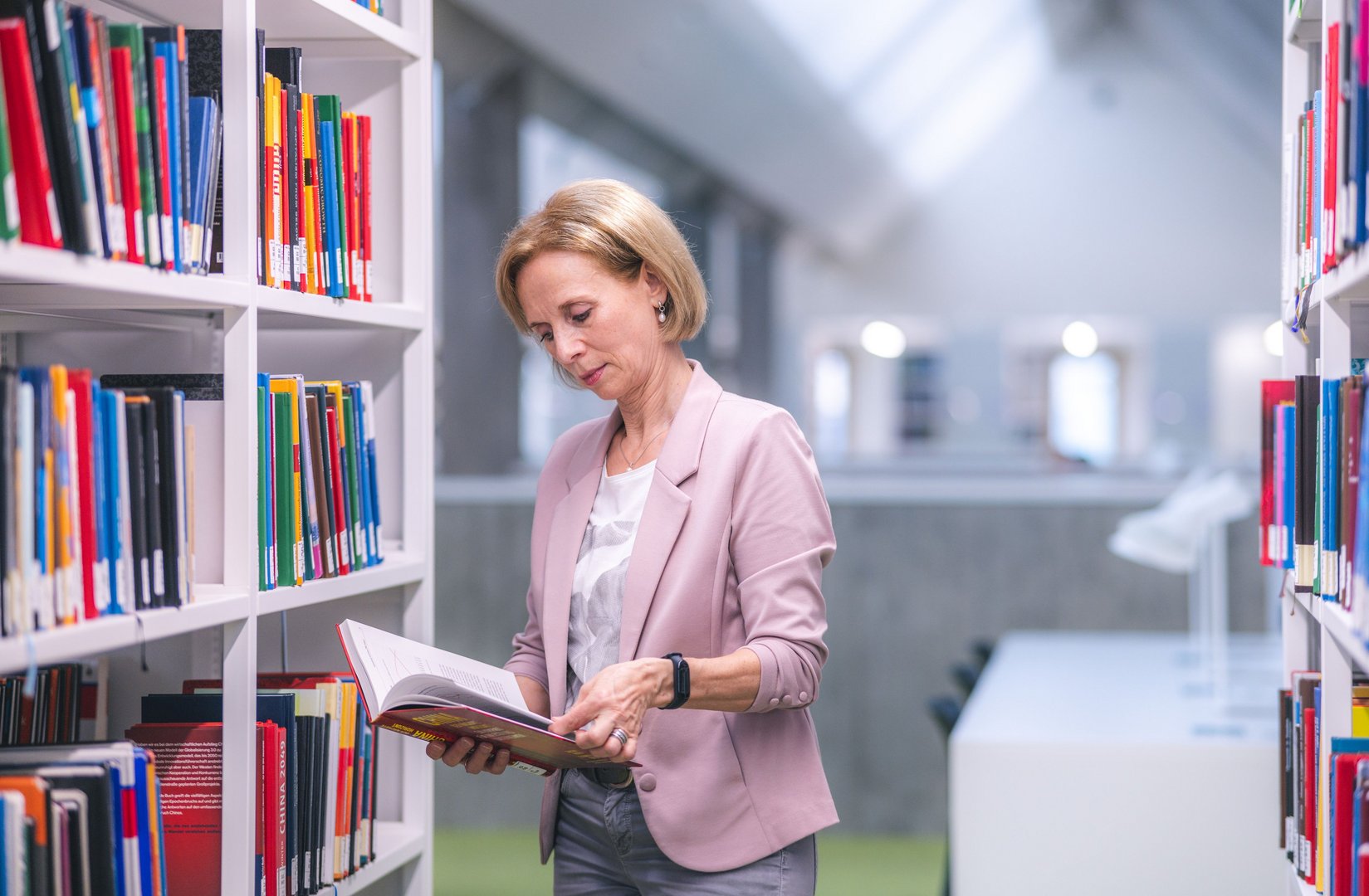 Eine Frau mit blonden Haaren blättert in einem Buch, während sie in einer modernen Bibliothek zwischen Regalen steht.