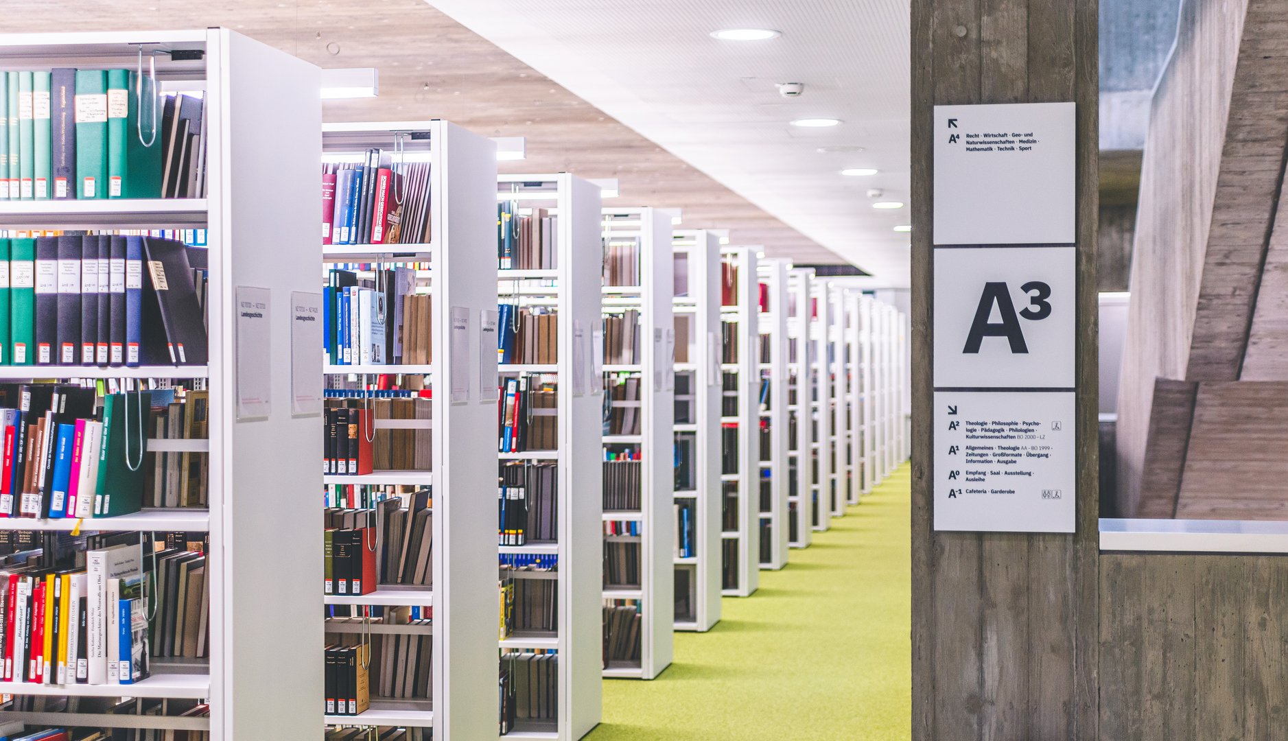 Ein moderner Bibliotheksraum mit hohen Regalen, die mit bunten Büchern und Ordnern gefüllt sind, auf einem grünen Teppichboden.
