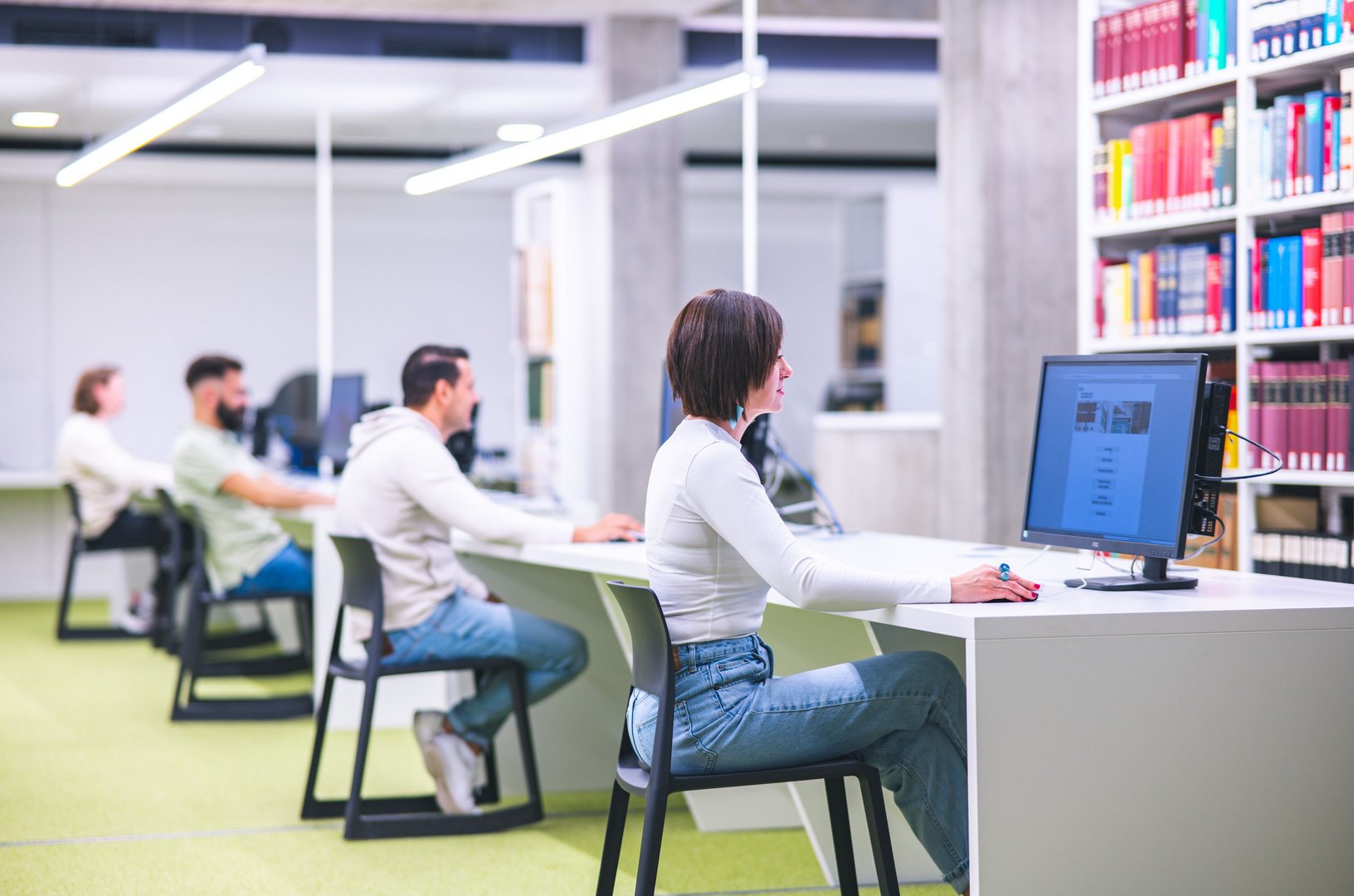 In einem modernen Raum arbeiten mehrere Personen an Computern, während Bücherregale im Hintergrund sichtbar sind.