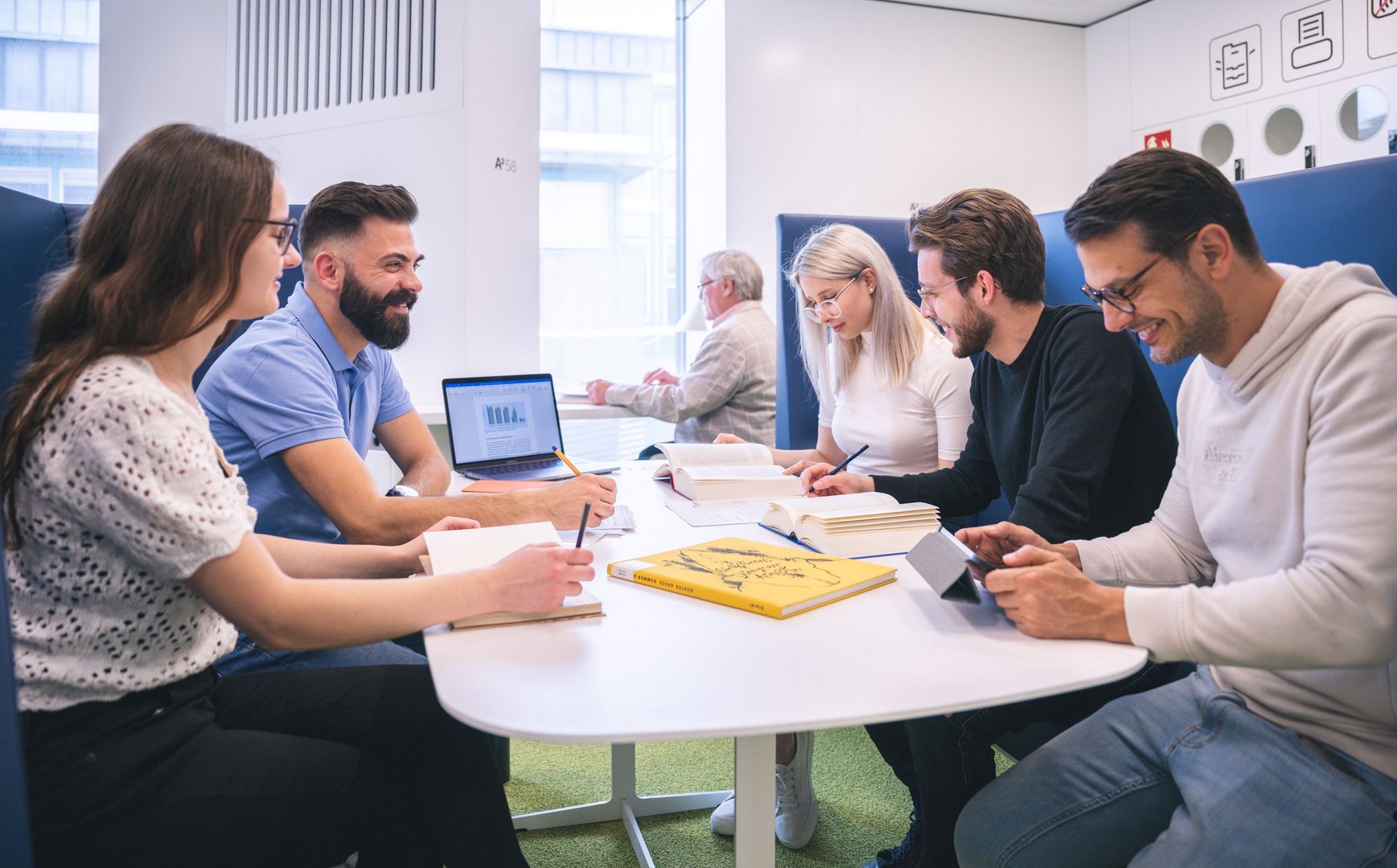 Eine Gruppe von Studierenden arbeitet gemeinsam an einem Tisch, umgeben von Büchern und Notizen, in einem modernen Raum.