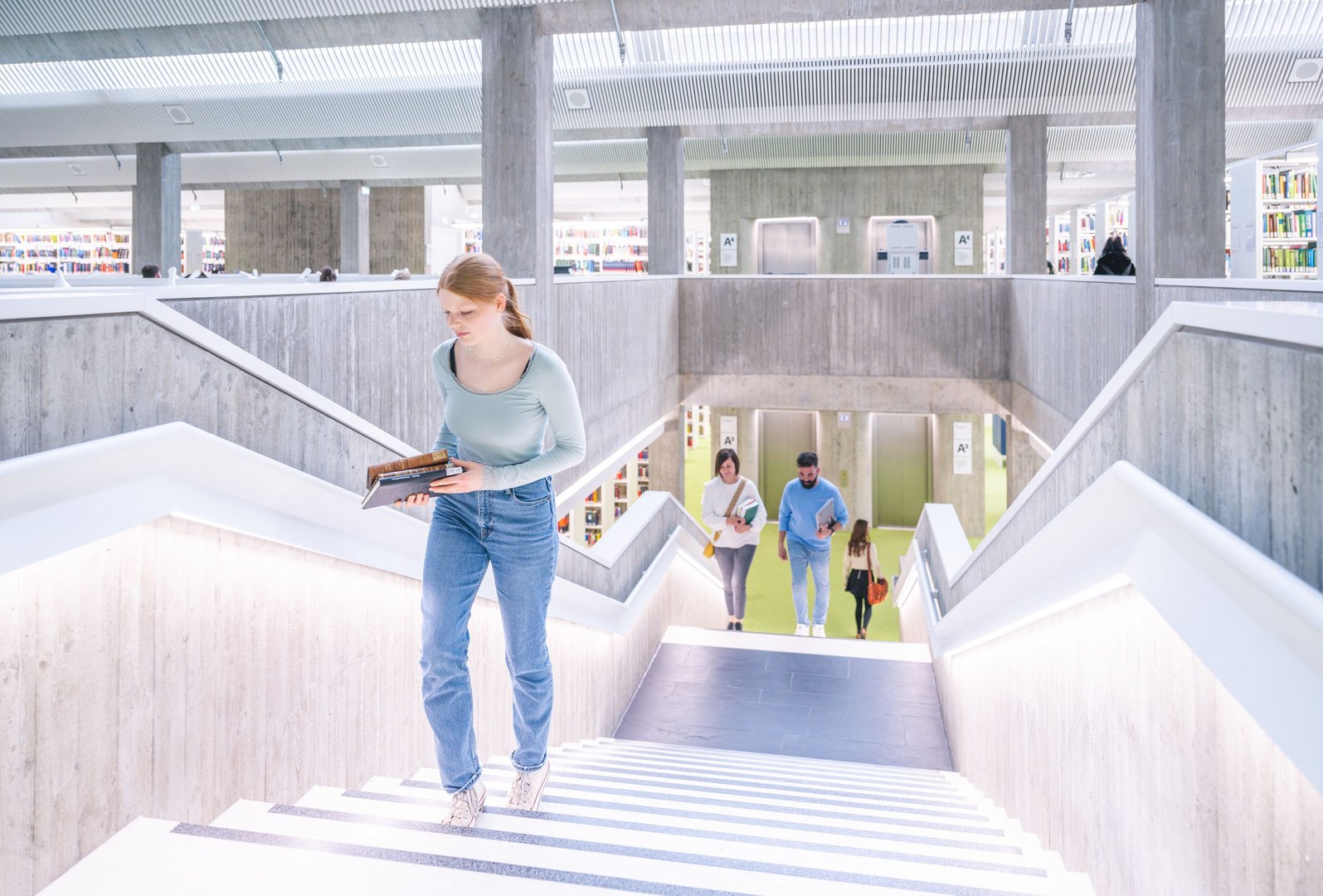 Eine junge Frau trägt ein Buch und geht die Treppe in der WLB hinauf.