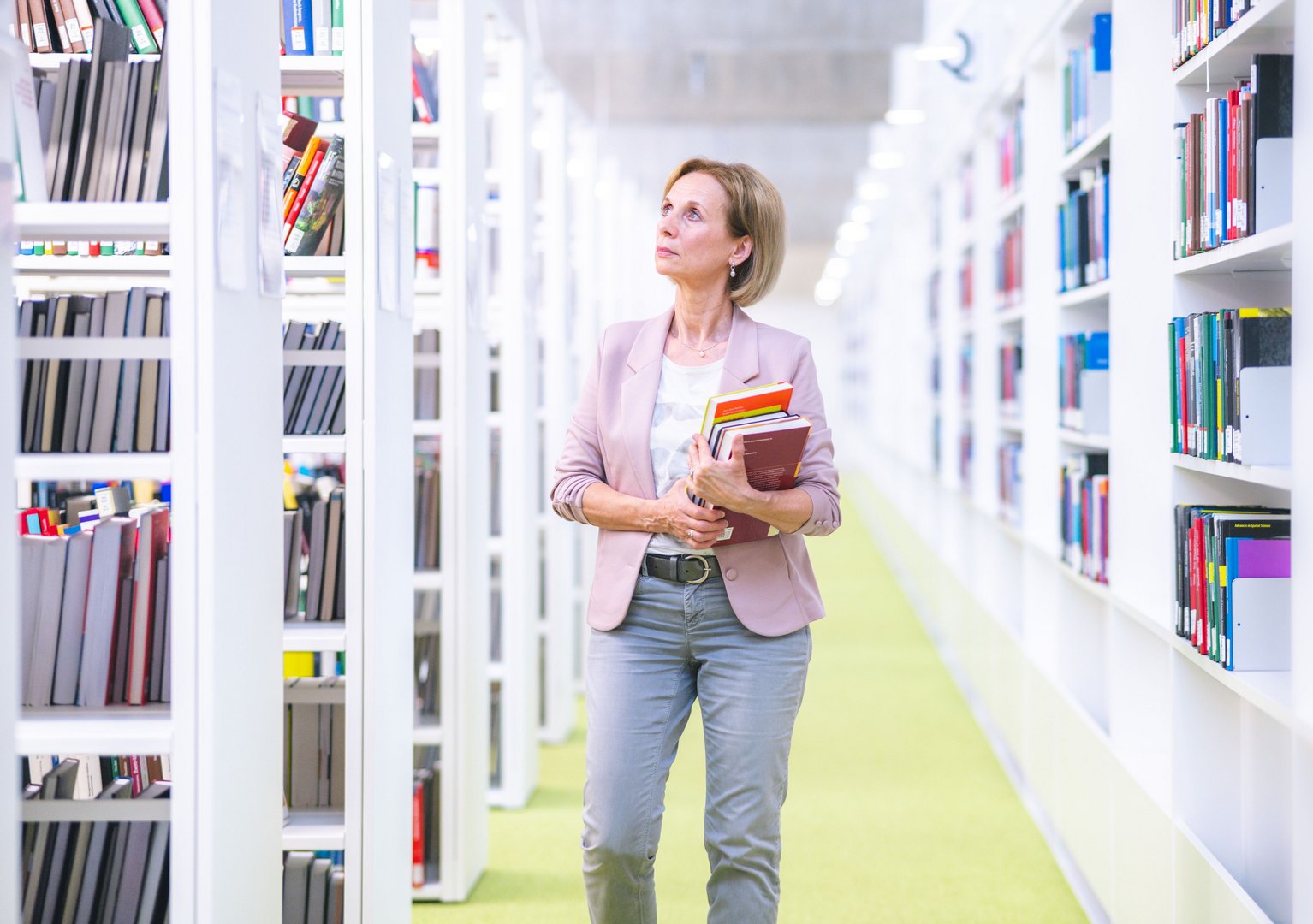 In einer hellen Bibliothek steht eine Frau mit einem Stapel Bücher in den Händen und betrachtet die Regale voller Literatur.