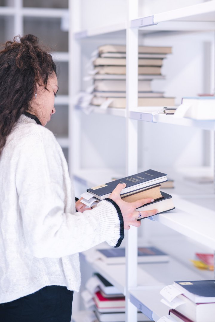 Eine Person hält mehrere Bücher in einer modernen Bibliothek und schaut auf die Regale.