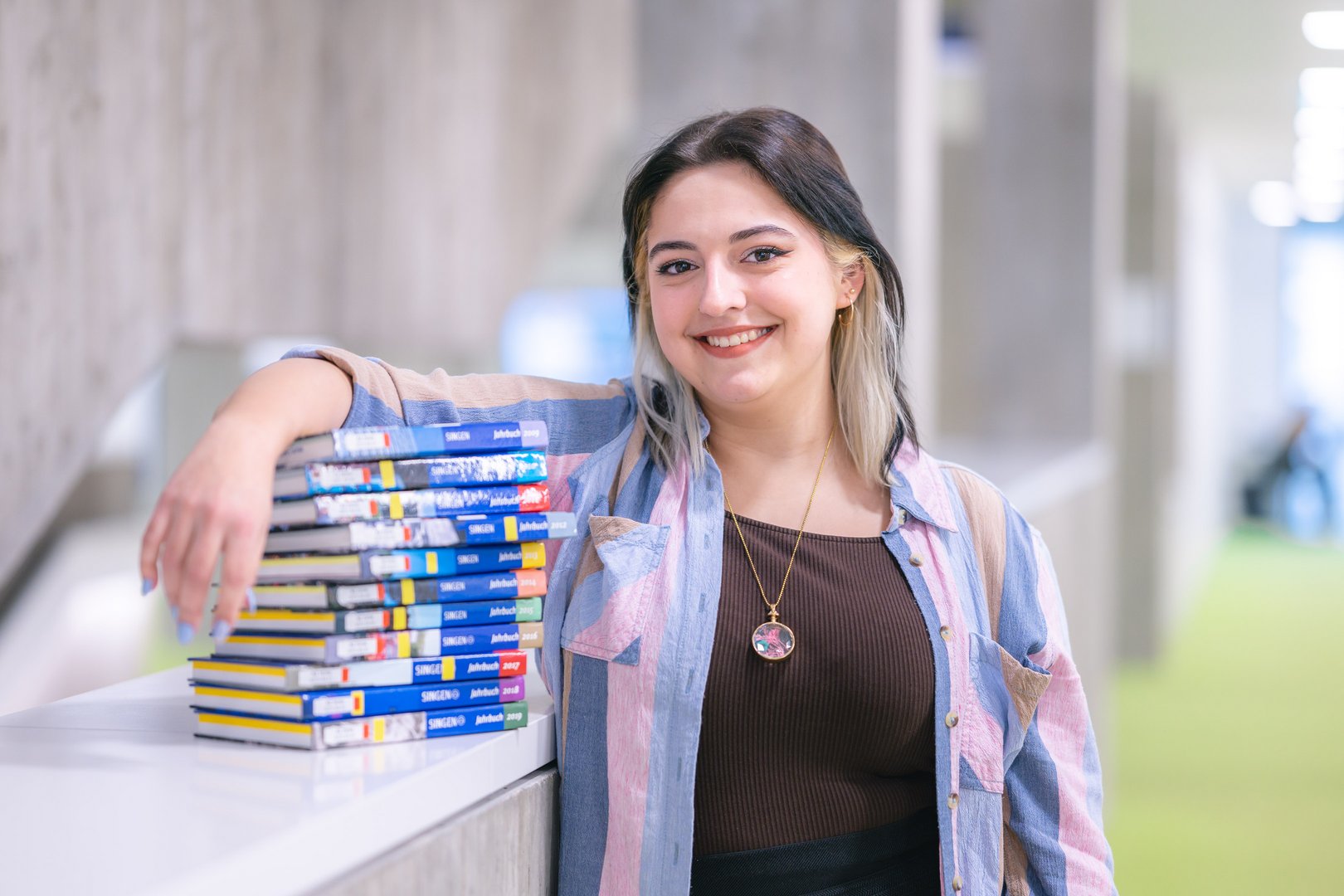 Eine junge Frau lächelt, während sie an einem Tisch mit einer Stapel von bunten Büchern lehnt.