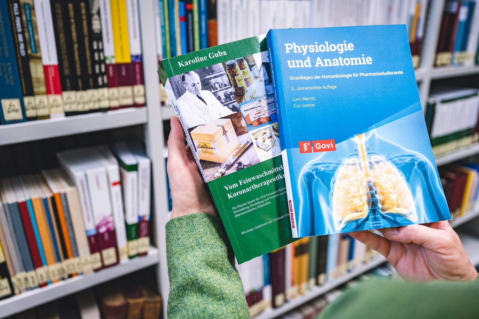 Eine Hand hält zwei Bücher in einer Bibliothek, eines über Physiologie und das andere über Koronartherapeutika.