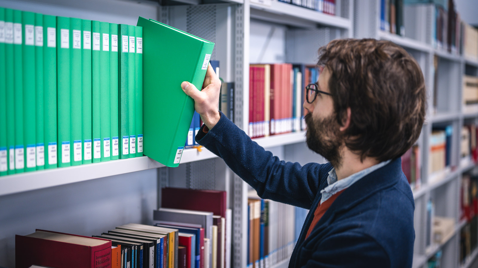 Ein Mann in einem blauen Blazer sucht in einem Regal nach einem grünen Buch zwischen vielen anderen Büchern.