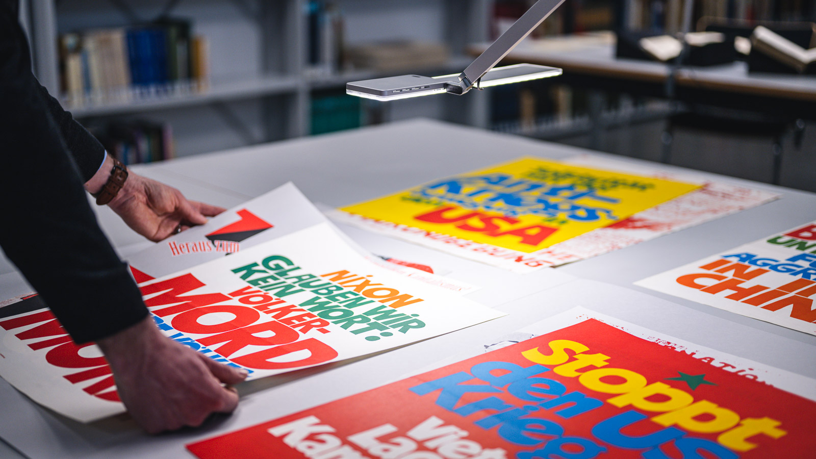 Protestplakate mit verschiedenen Botschaften liegen auf einem Tisch, während eine Hand ein Plakat mit 'MORD' anhebt.