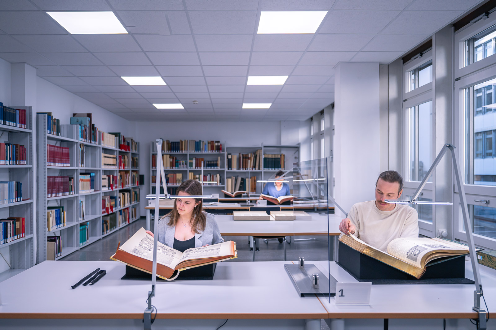 In einem modernen Lesesaal studieren drei Personen konzentriert alte Bücher, umgeben von Regalen voller Literatur.