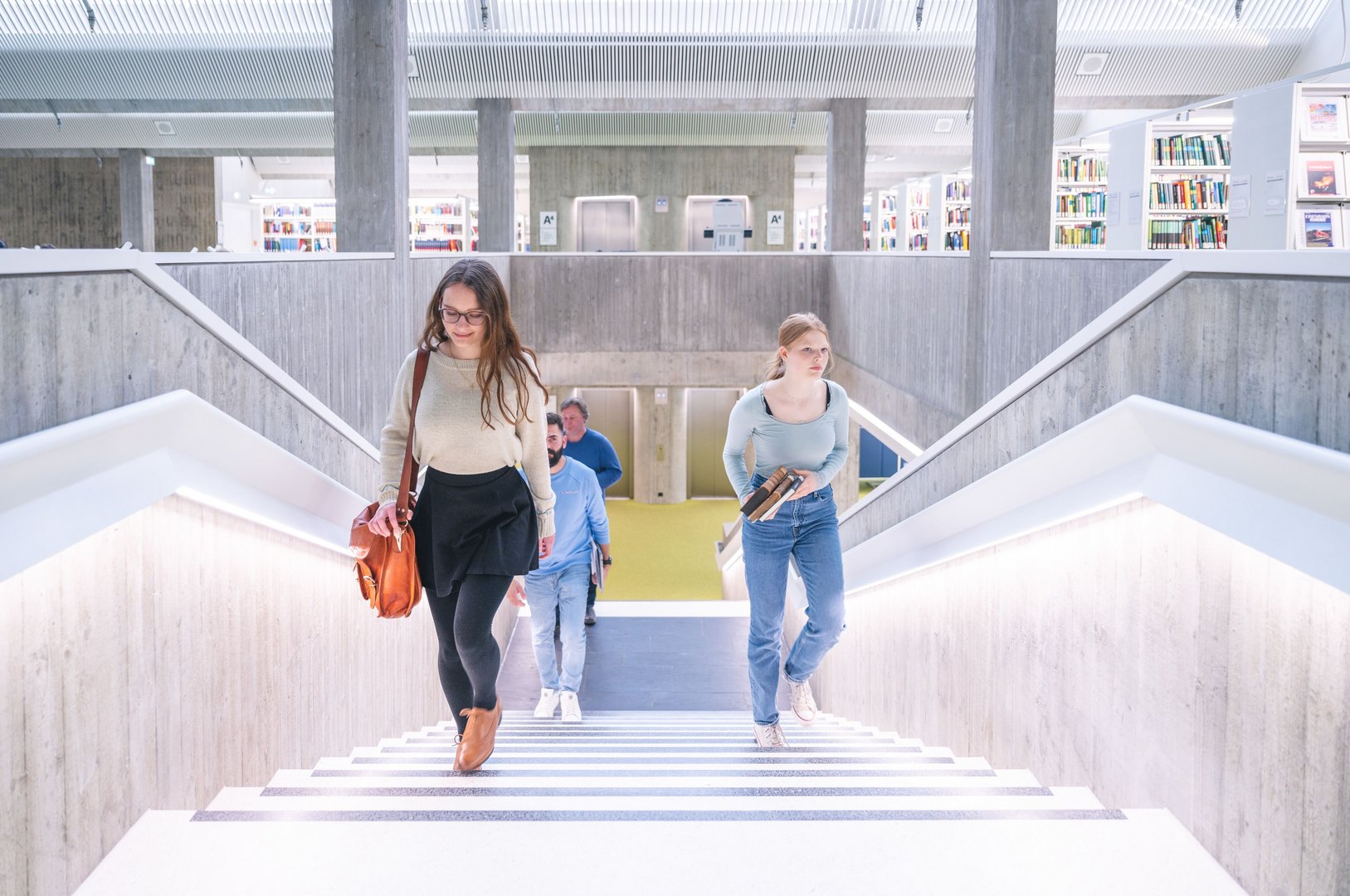 Drei Personen gehen die Treppe in einer modernen Bibliothek hinauf, umgeben von hellen Regalen voller Bücher.