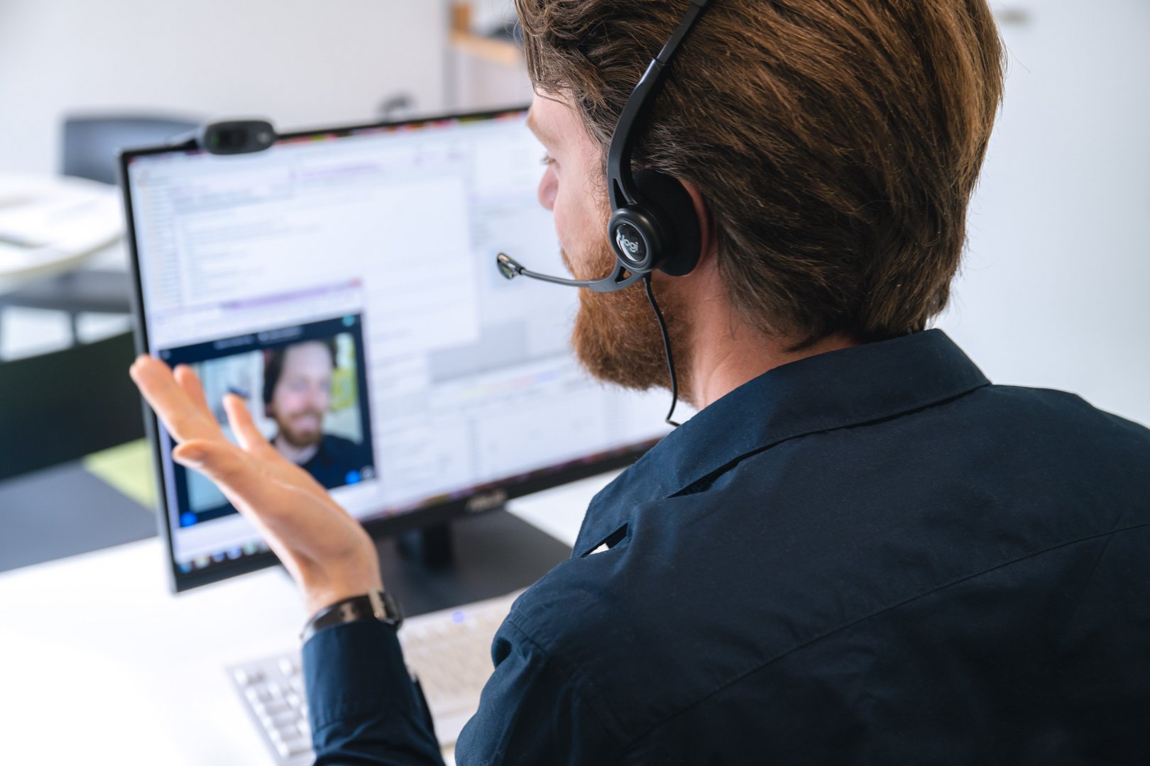 Ein Mann mit Headset spricht während eines Videoanrufs, seine Hand gestikuliert lebhaft in einem modernen Büro.
