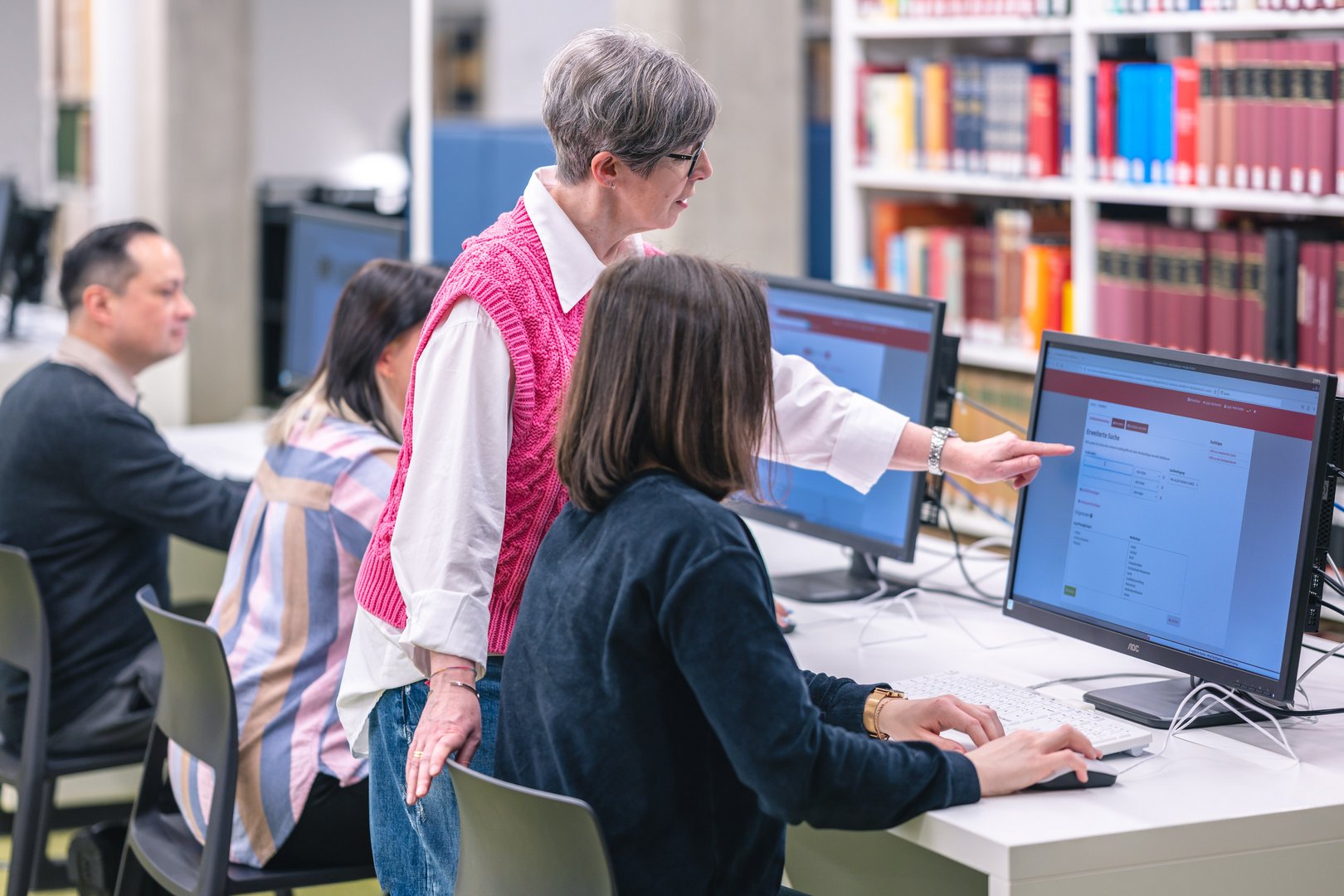 Eine ältere Frau erklärt einer jüngeren Person am Computer etwas, während andere Nutzer im Hintergrund arbeiten.