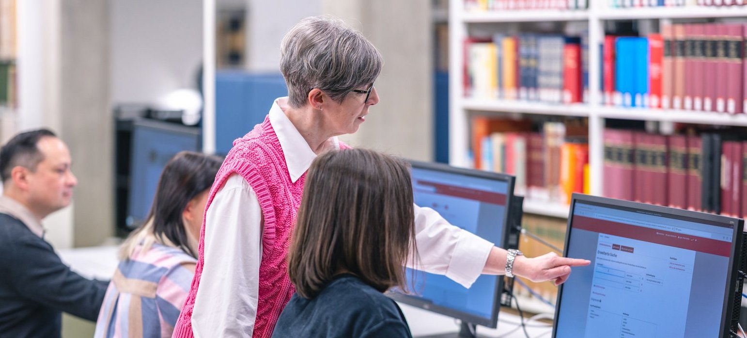 Eine Gruppe von Menschen sitzt an Computern, während eine Frau in einem rosa Pullover Anweisungen gibt und auf den Bildschirm zeigt.