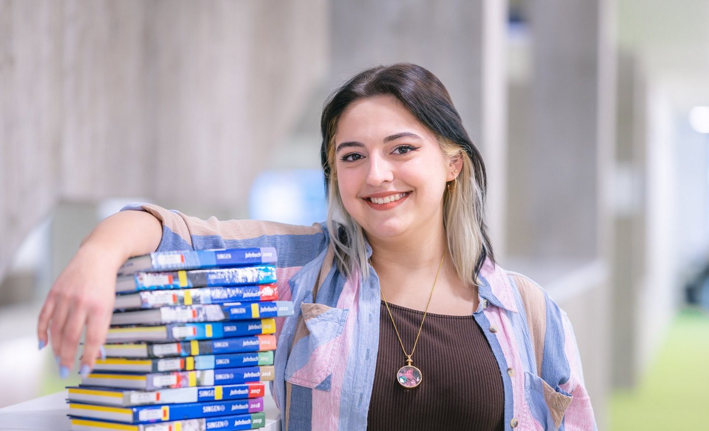 Eine junge Frau lächelt, während sie an einem Tisch mit einer Stapel von bunten Büchern lehnt.