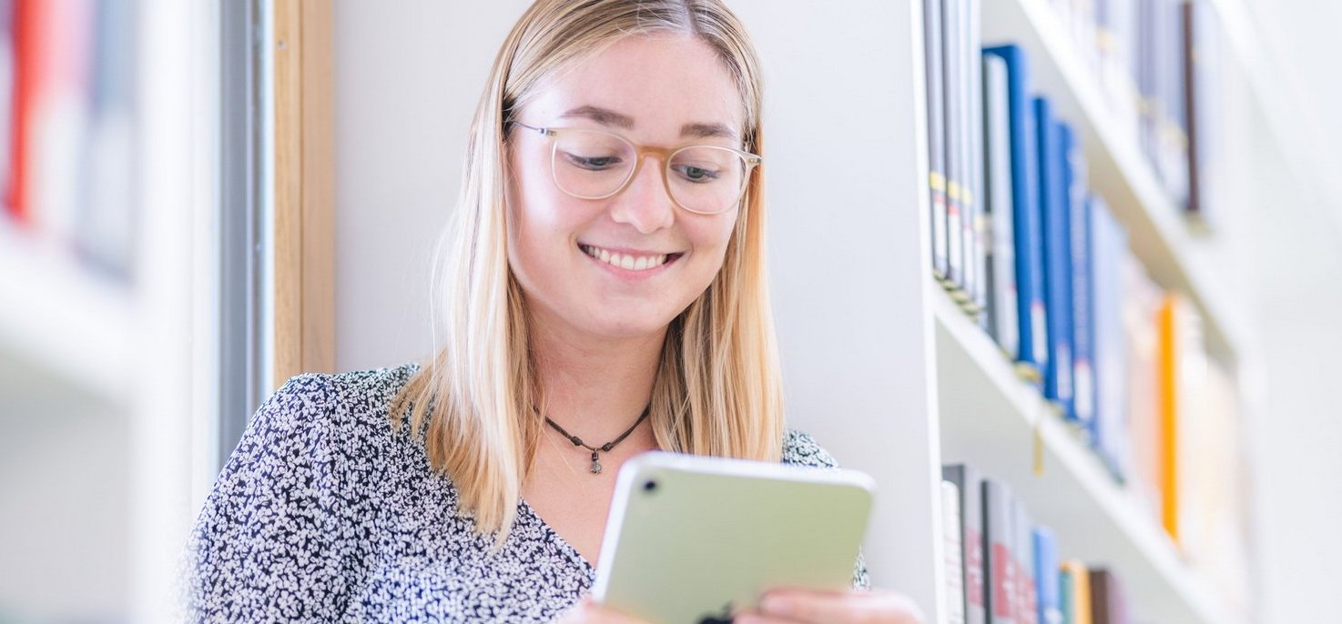 Eine junge Frau mit blonden Haaren lächelt, während sie ein Tablet in einer Bibliothek benutzt, umgeben von Bücherregalen.