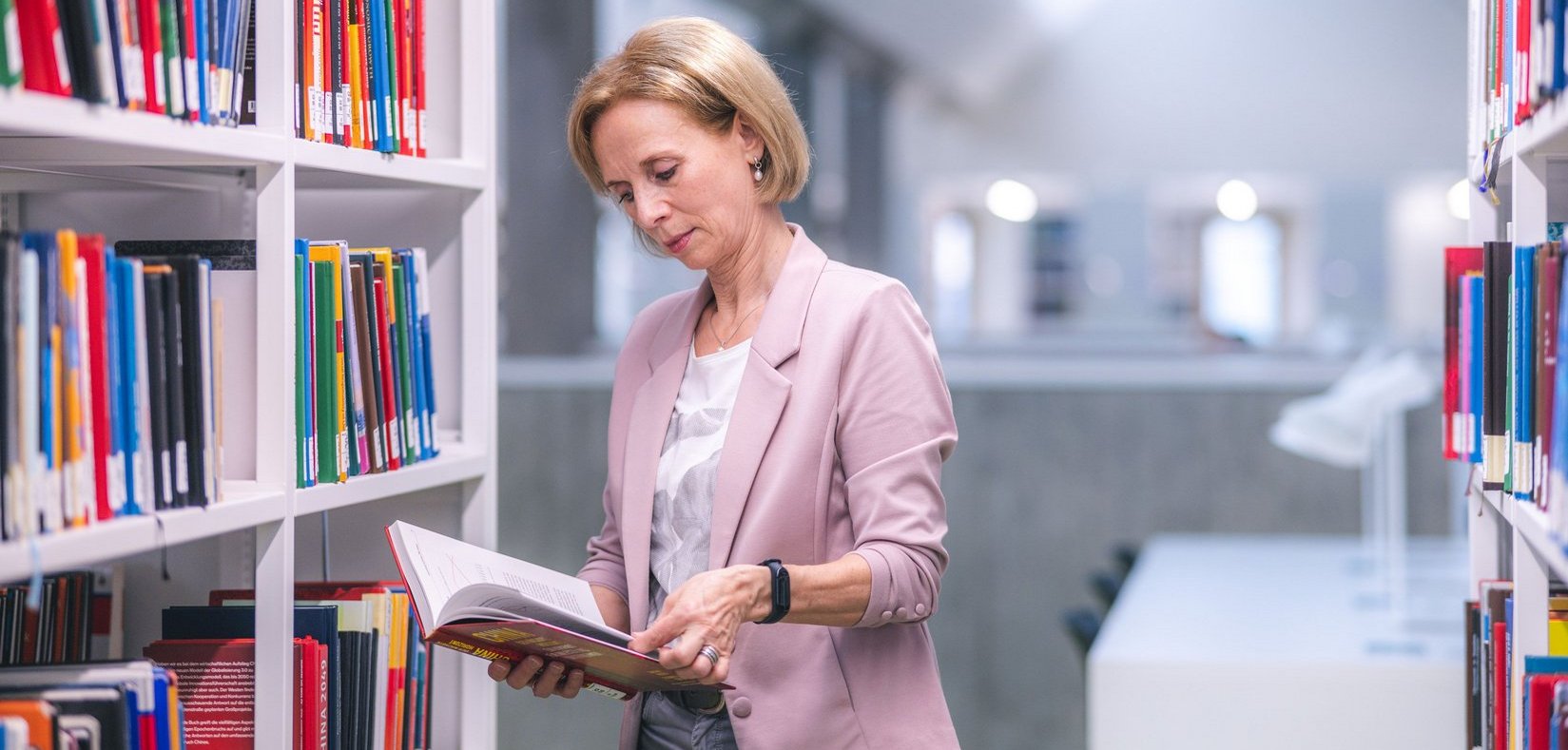 Eine Frau mit blonden Haaren blättert in einem Buch, während sie in einer modernen Bibliothek zwischen Regalen steht.