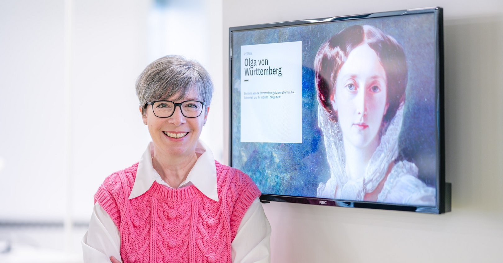 Eine lächelnde Frau mit grauen Haaren trägt einen pinkfarbenen Pullover und steht vor einem Bildschirm mit einem Porträt von Olga von Württemberg.